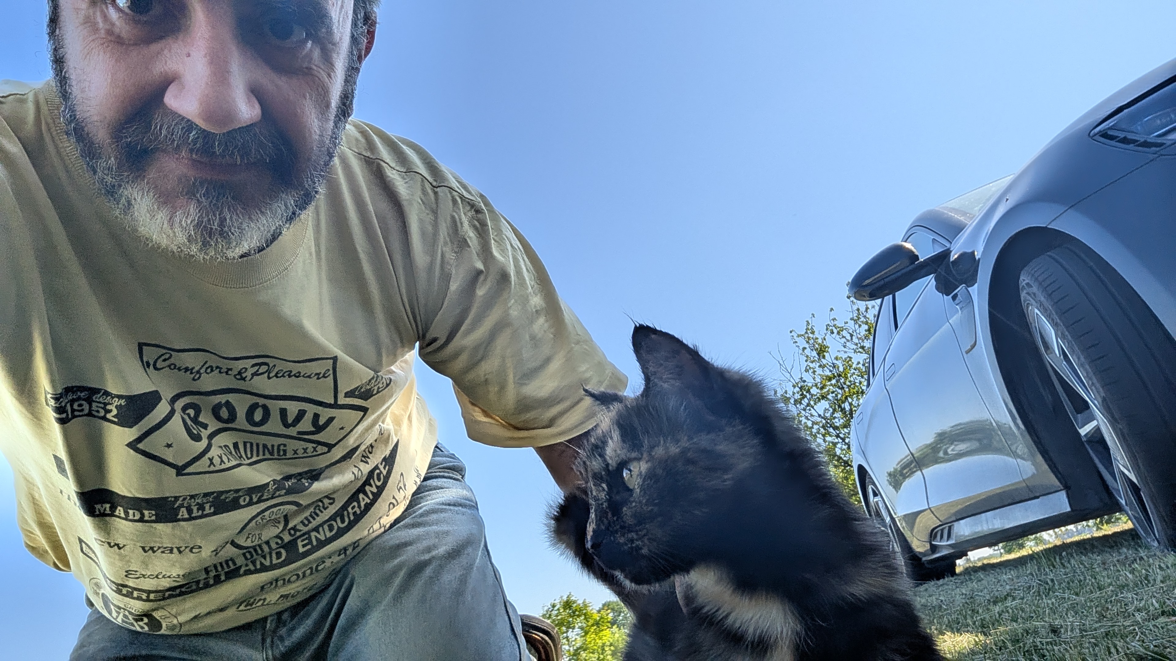 Low outdoor selfie of Andrea with a tortoiseshell cat; grass, parked car, and blue sky