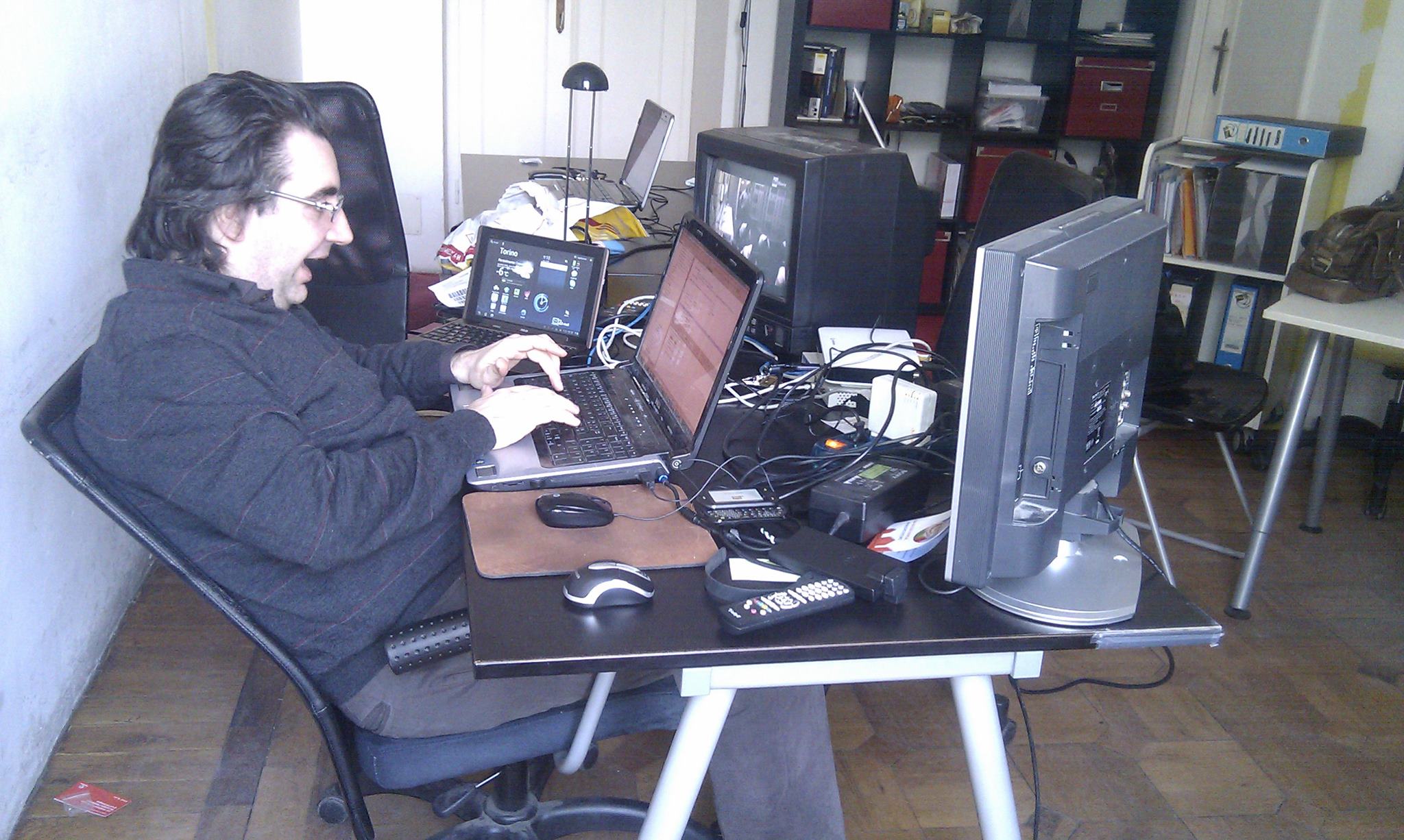 Andrea at a cluttered desk with a laptop, a small netbook showing a Torino weather widget, a CRT and a flat-panel monitor, mice, remotes, and tangled cables on parquet