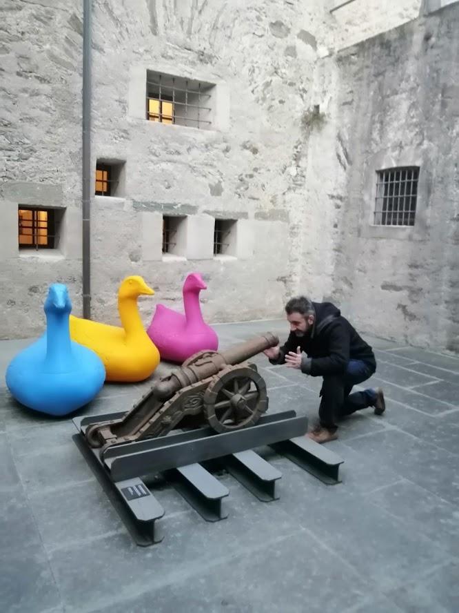 The Poltro kneeling behind an antique cannon aimed at three giant colourful duck sculptures in a castle courtyard