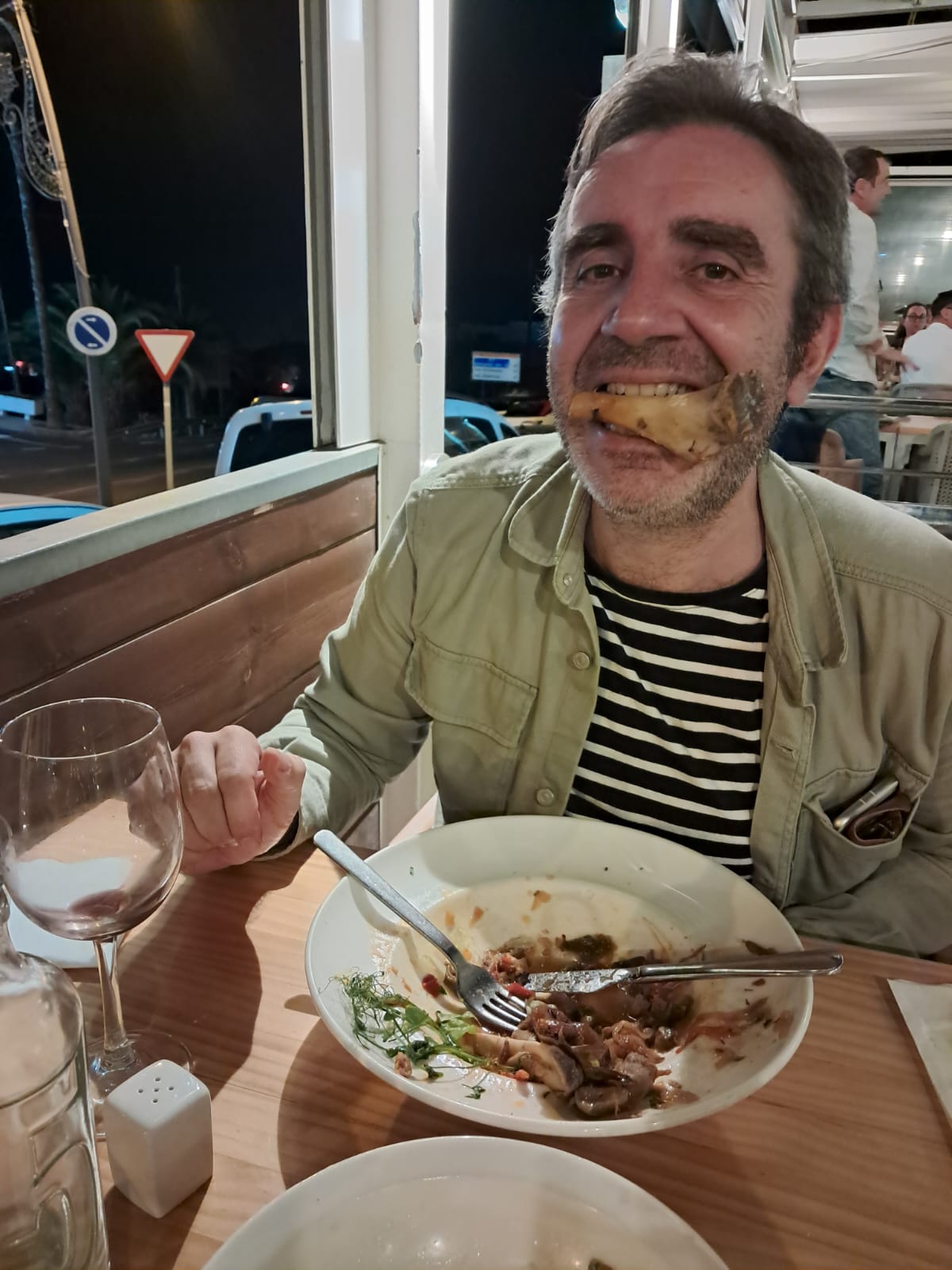 The Poltro grinning at a restaurant table with a bone from the boar dish clenched between his teeth like a tusk, an empty plate in front of him