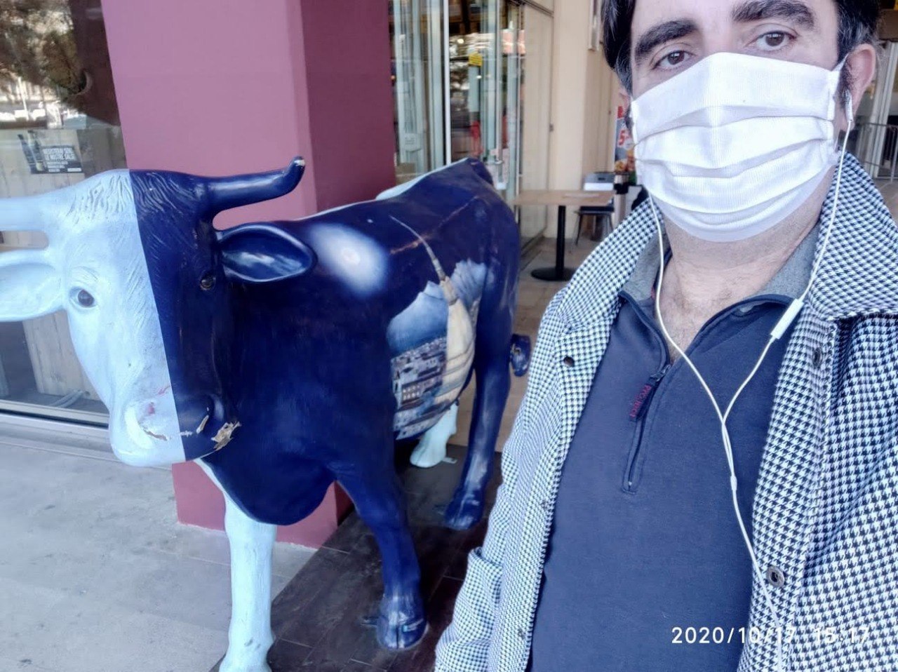 The Poltro in a surgical mask taking a selfie next to a painted fibreglass cow outside a shop, timestamp in the corner