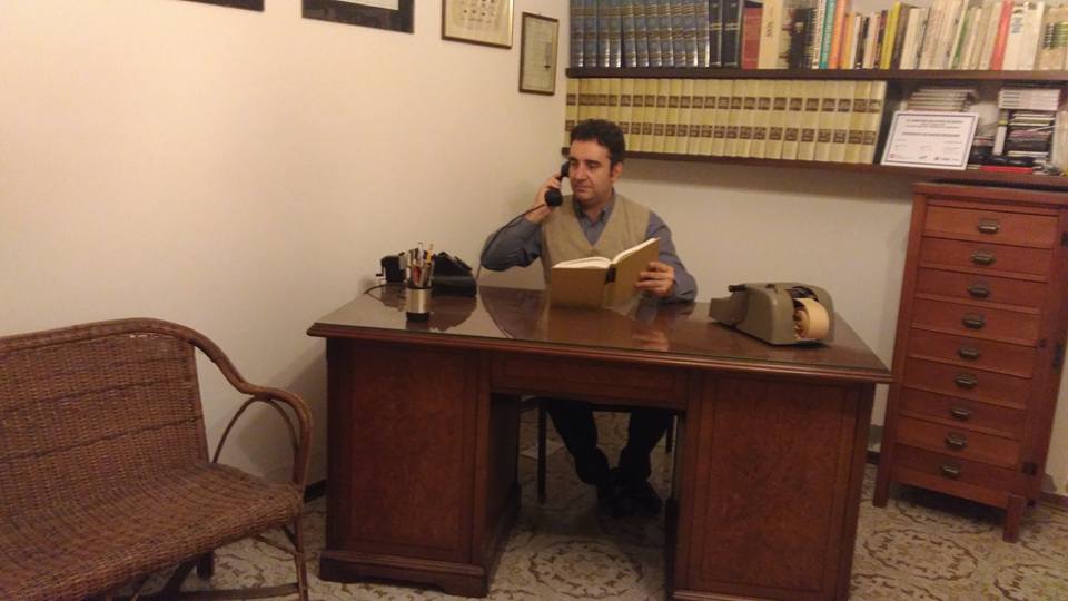Andrea at a large wood desk on the phone, reading a book, with rotary telephone and mechanical adding machine; bookshelves behind