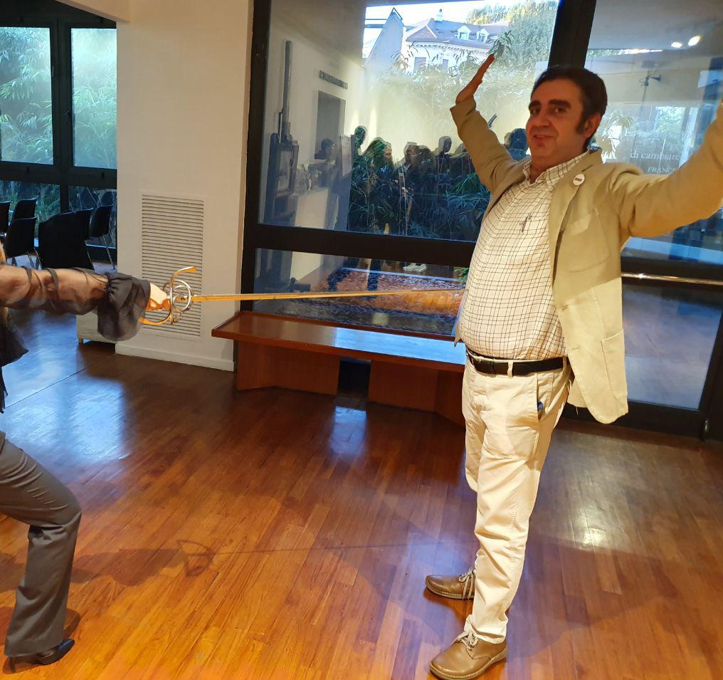 Andrea smiling with hands raised in mock surrender while someone points a rapier toward him; parquet floor and tall windows